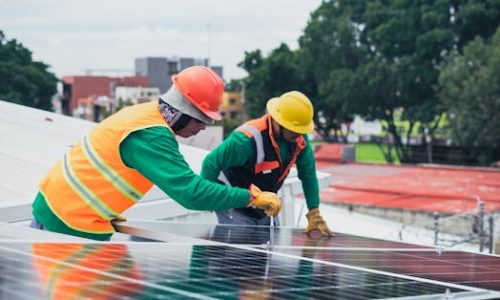 Two workers installing rooftop solar panels safely equipped with PPE and tools.